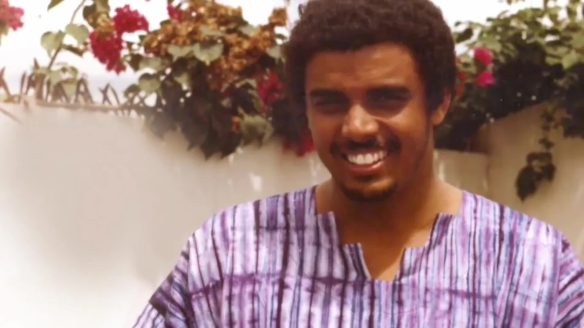 A young Dag Heward-Mills smiling in a purple striped smock, bougainvillea behind him.