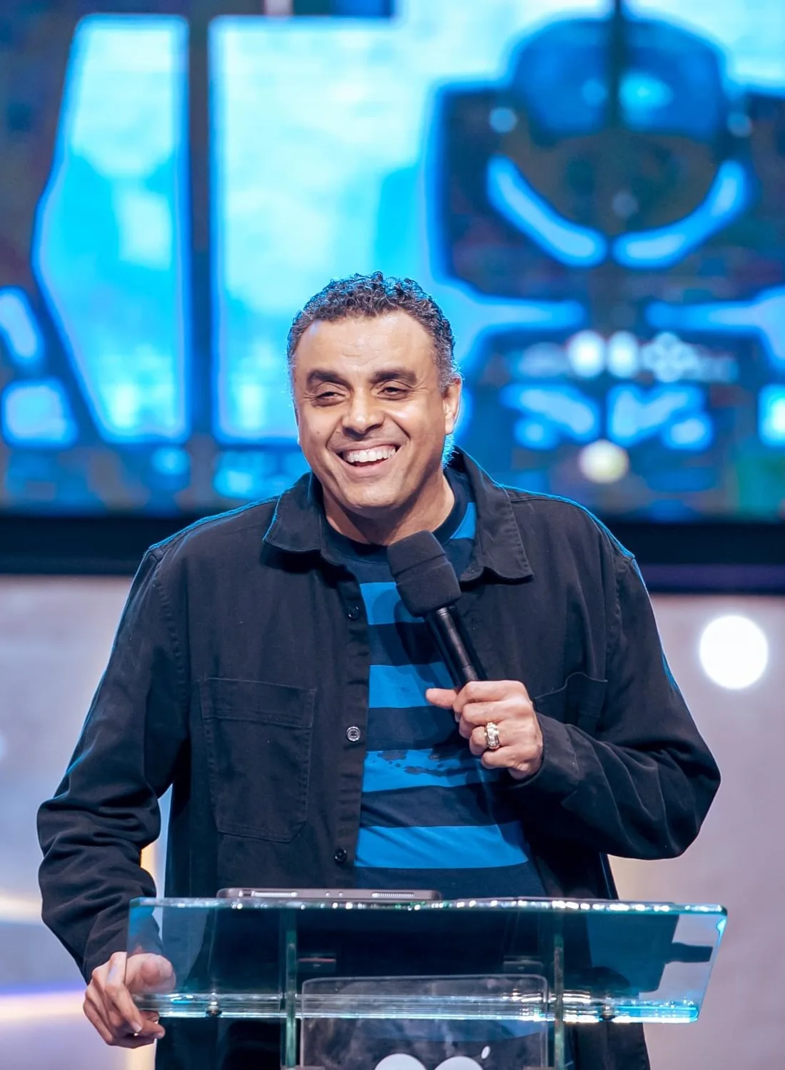 Bishop Dag Heward-Mills lacht breed aan een glazen preekstoel, met een microfoon in de hand.